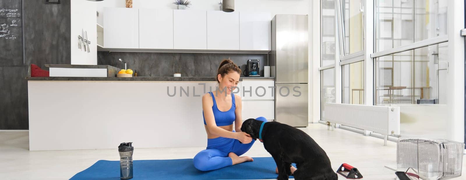 Portrait of beautiful young woman tries to do fitness workout from home, sits on floor on yoga mat with her cute dog, puppy wants to play while girl does exercises.