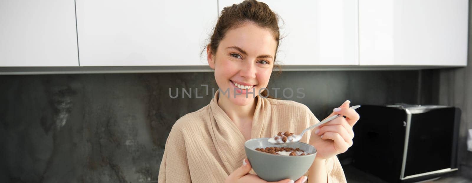 Gorgeous woman starts her day with cereals, eats breakfast and smiles, wears bathrobe, leans on kitchen worktop, starts her morning with quick meal.
