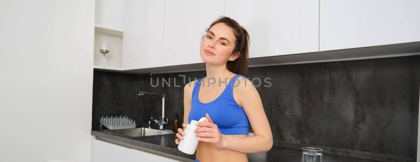 Image of young brunette woman with slim, fit body, holding dietary supplements, vitamin B, fish oil, standing in kitchen.