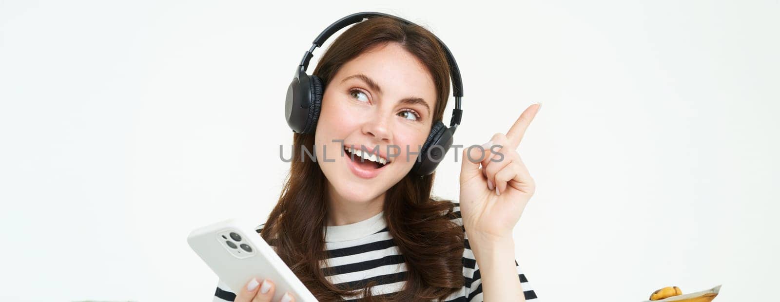 Happy beautiful woman, listening music in wireless headphones, holding mobile phone, pointing left at copy space, showing advertisement, white background.