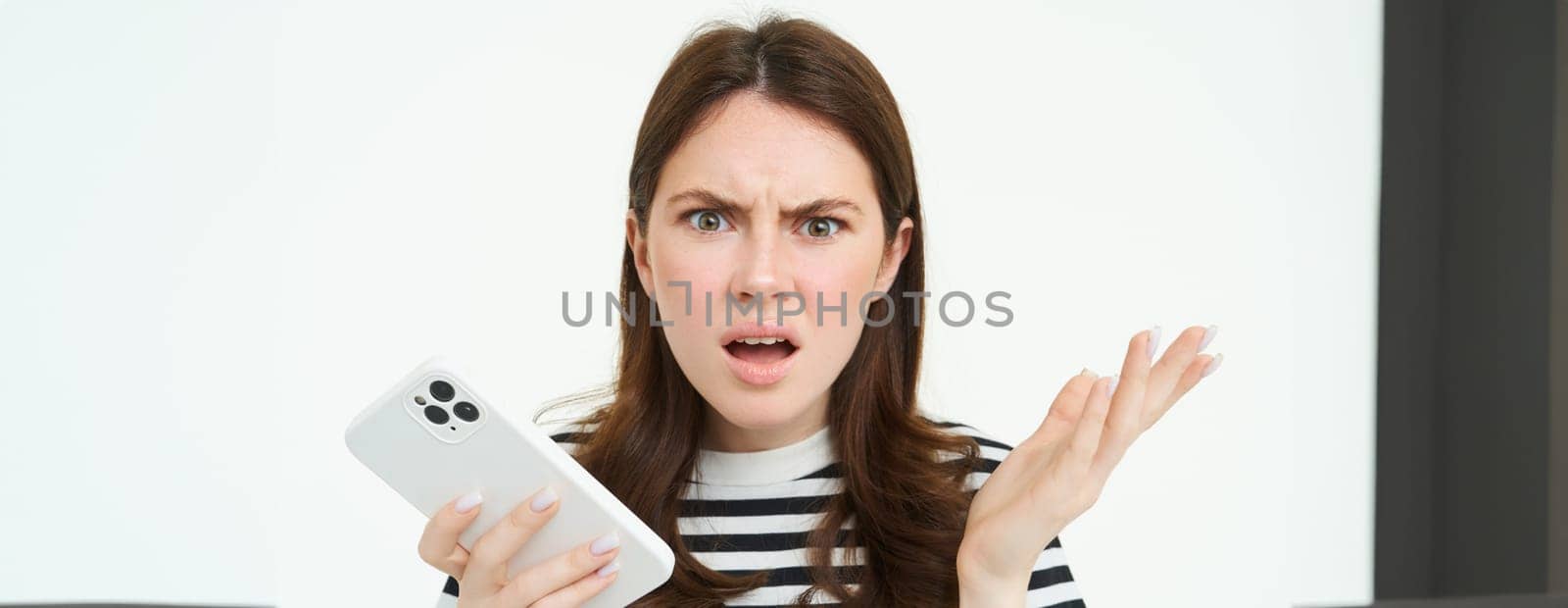 Portrait of angry, confused young woman shrugging shoulders while using mobile phone, holding smartphone with annoyed face expression, frowning, standing over white background.