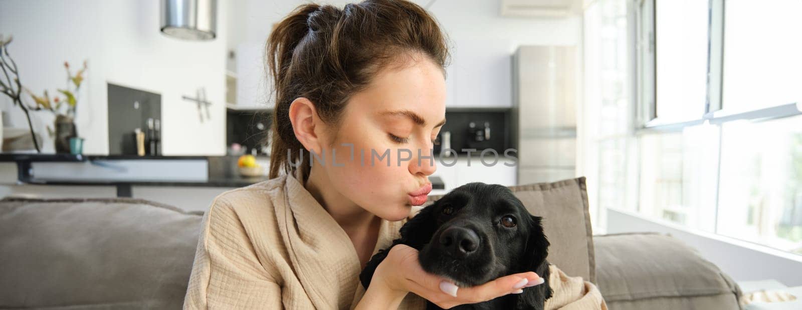 Portrait of young woman kissing her cute dog, cuddling her four-legged friend on sofa, spending time with puppy.
