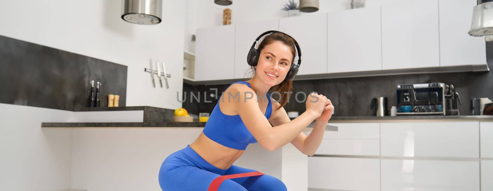 Portrait of active and healthy young woman, doing squats with resistance band, workout at home, listening music in wireless headphones.