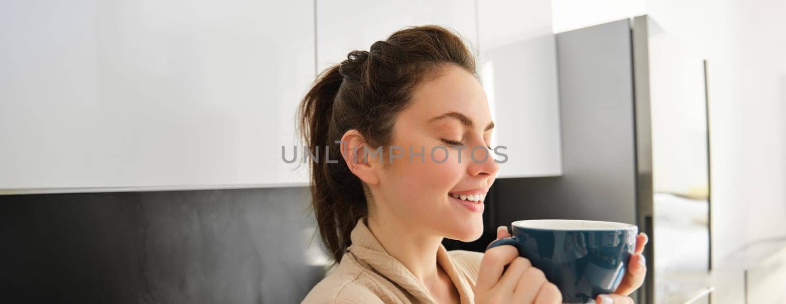 Portrait of happy young woman starts her morning with mug of coffee, drinking tea from cup, standing in the kitchen, smiling cheerfully by Benzoix