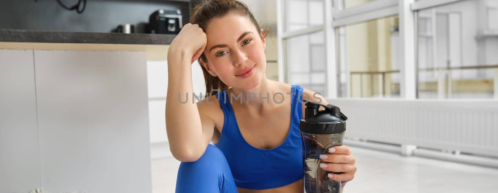 Beautiful young sportswoman drinks water, stays hydrated during workout training at home, doing fitness exercises in living room.