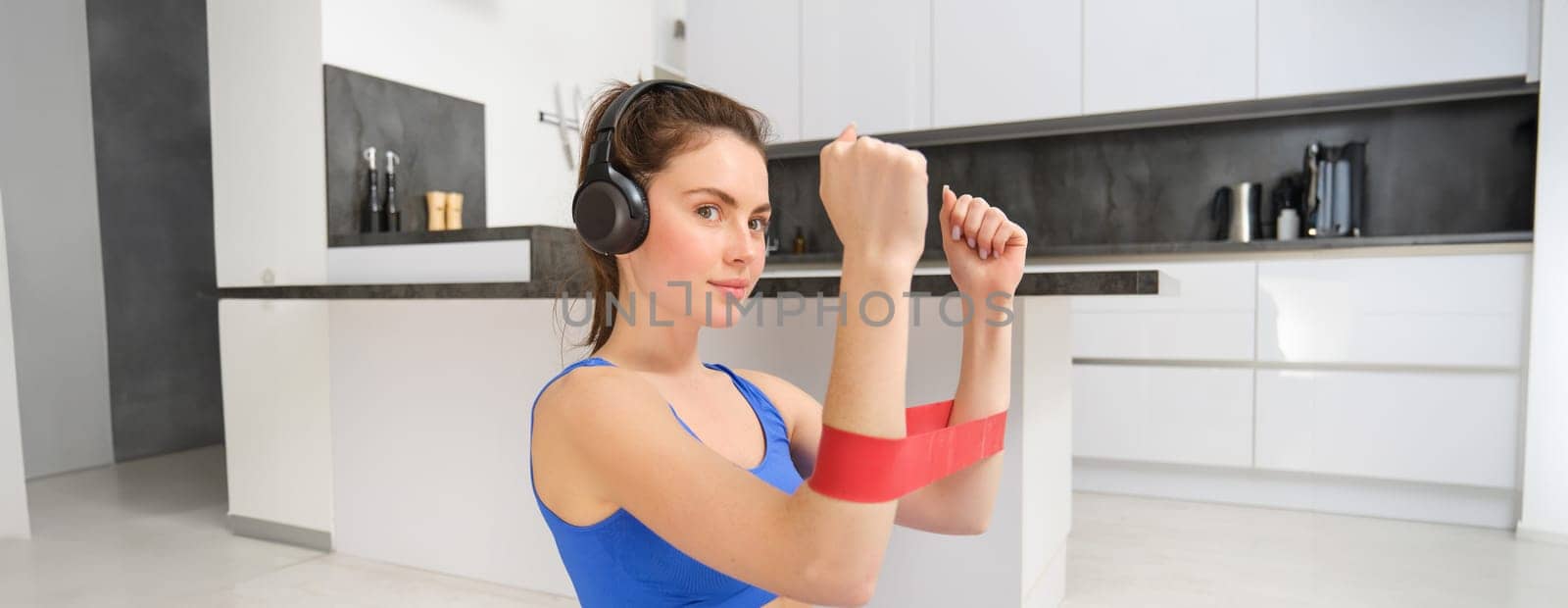 Young woman doing fitness workout at home, using elastic resistance band on arms, muscle exercises, sitting on rubber mat in living room.