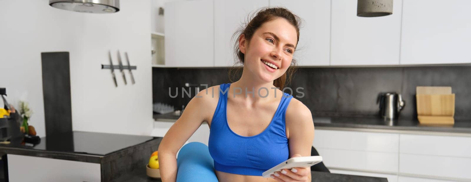 Portrait of fit and healthy woman doing workout at home, holding smartphone and rubber mat, smiling.