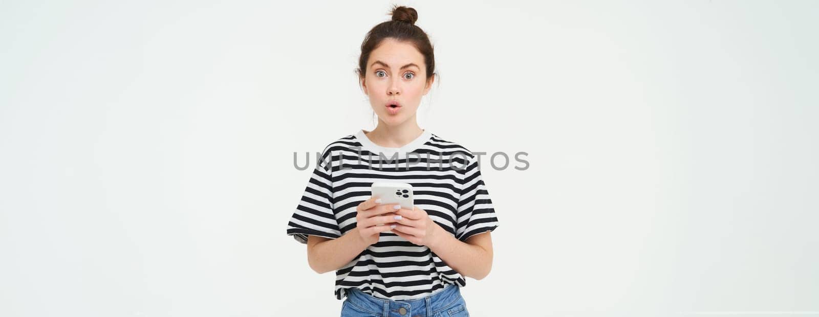 Portrait of impressed girl looks amazed, gasping, holding mobile phone, standing against white background by Benzoix