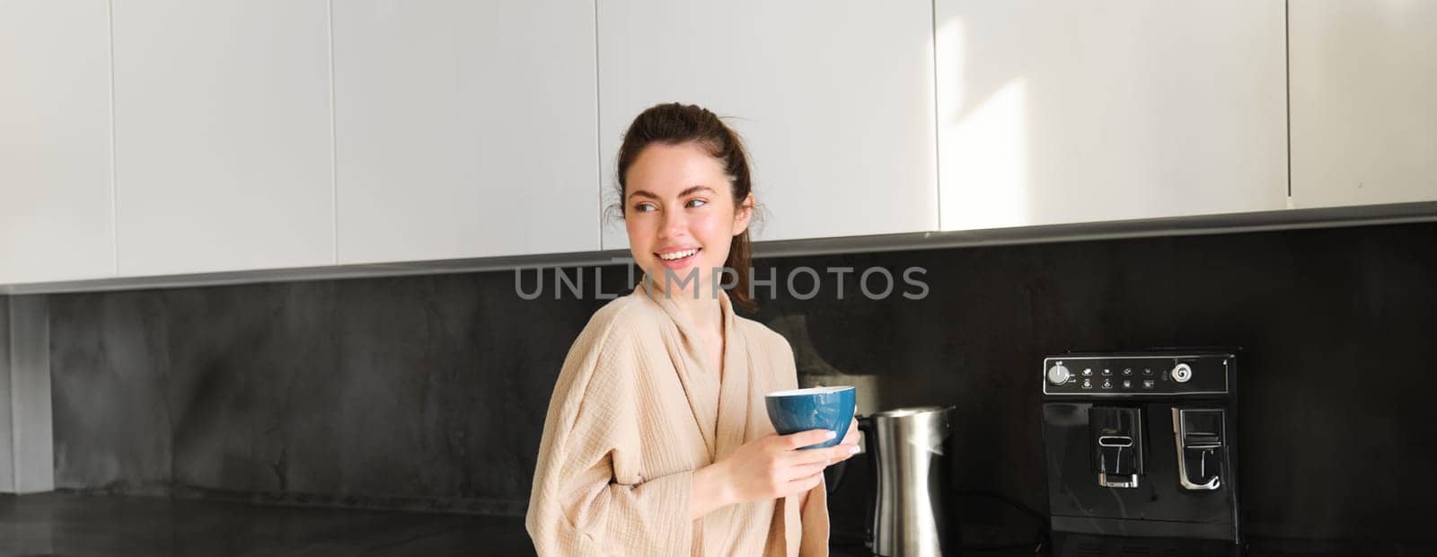 Portrait of beautiful housewife, young woman in bathrobe, holding cup of coffee, drinking tea in the kitchen, enjoying morning.