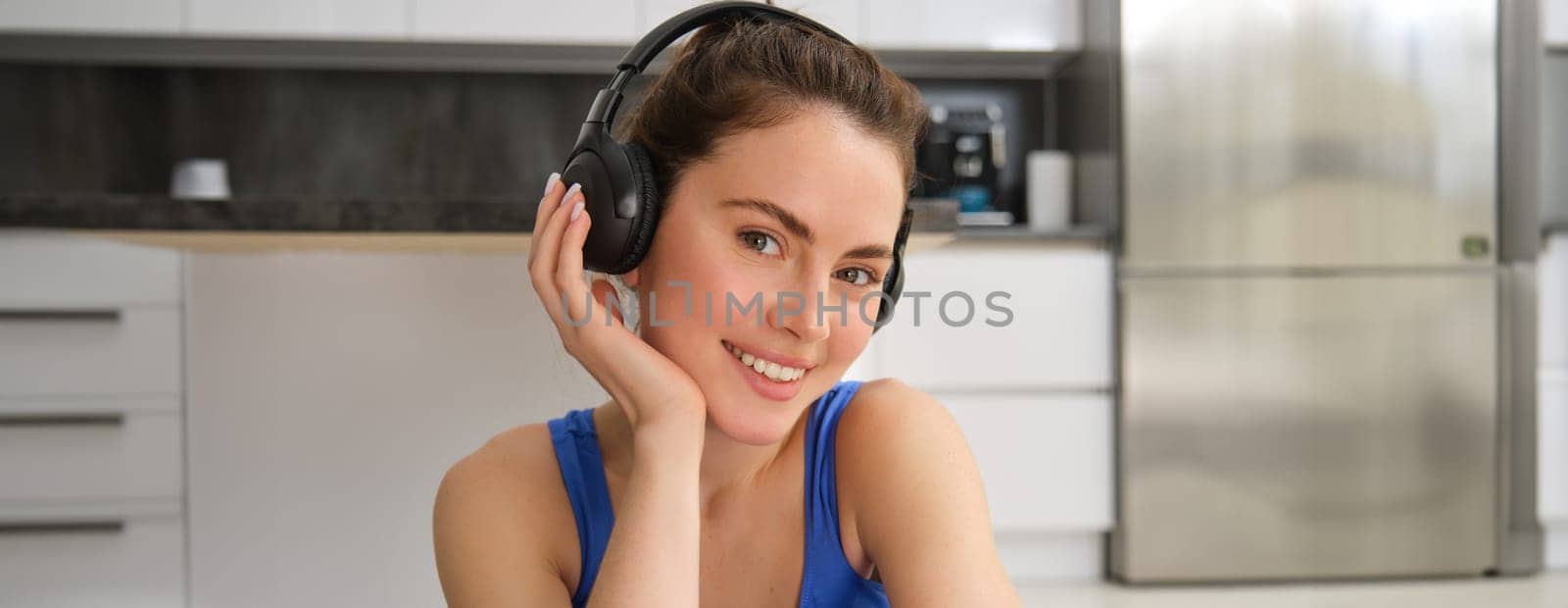 Close up portrait of beautiful brunette girl in sportswear, sits in wireless headphones and listens music, while doing workout training at home by Benzoix