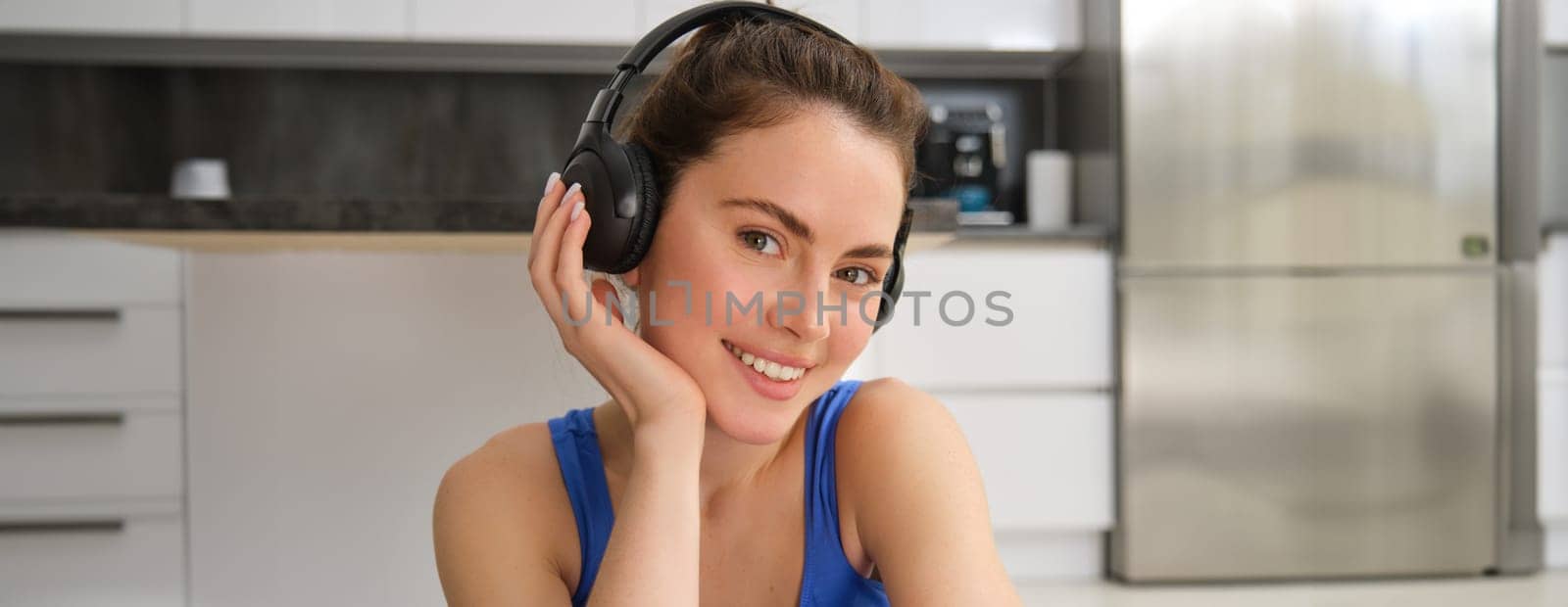 Close up portrait of beautiful brunette girl in sportswear, sits in wireless headphones and listens music, while doing workout training at home.