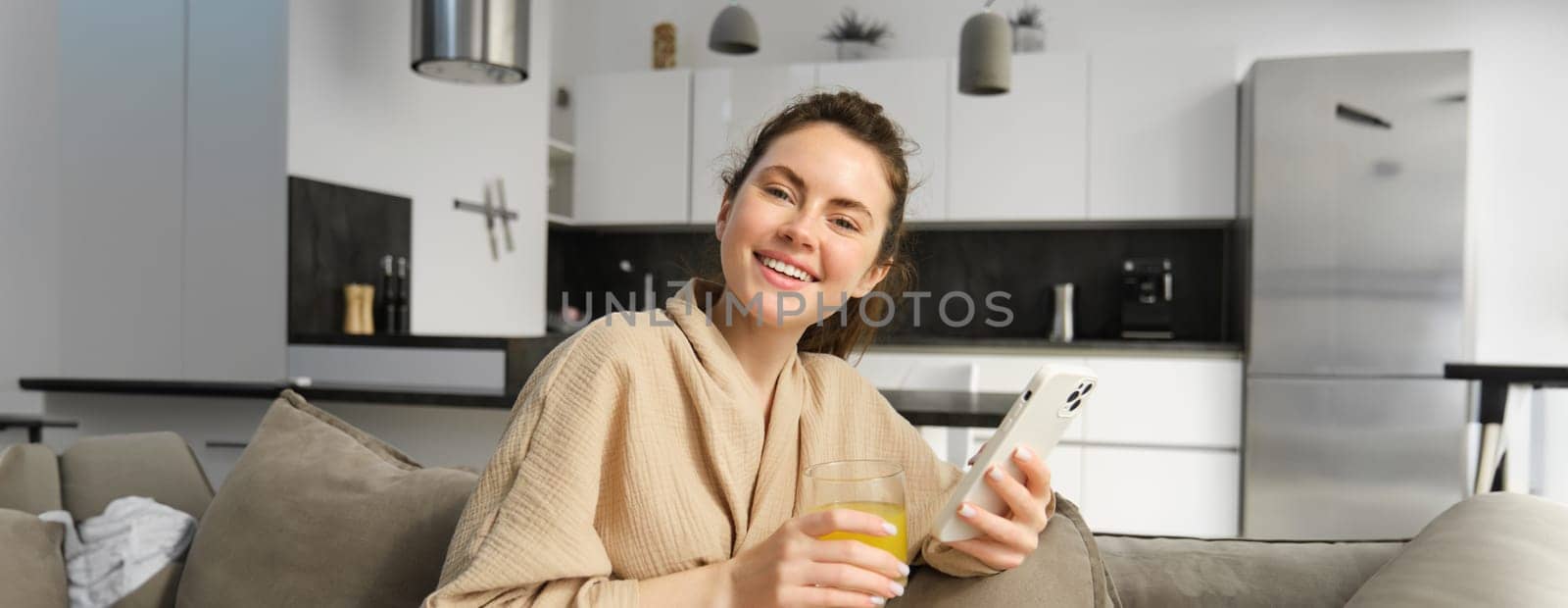 Beautiful girl in bathrobe, resting on couch at home, using smartphone, browsing social media online on mobile phone, drinking orange juice.