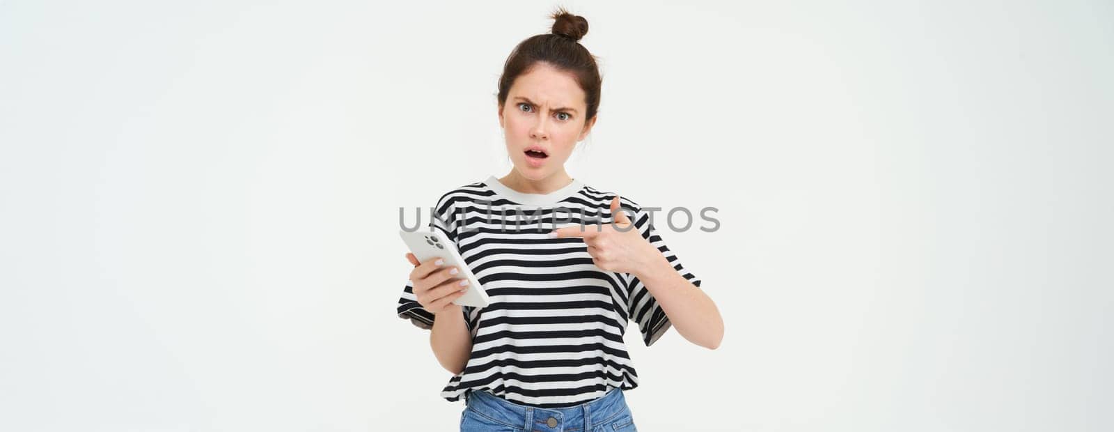 Image of angry young woman complains, points at telephone and looks disappointed, isolated against white background by Benzoix
