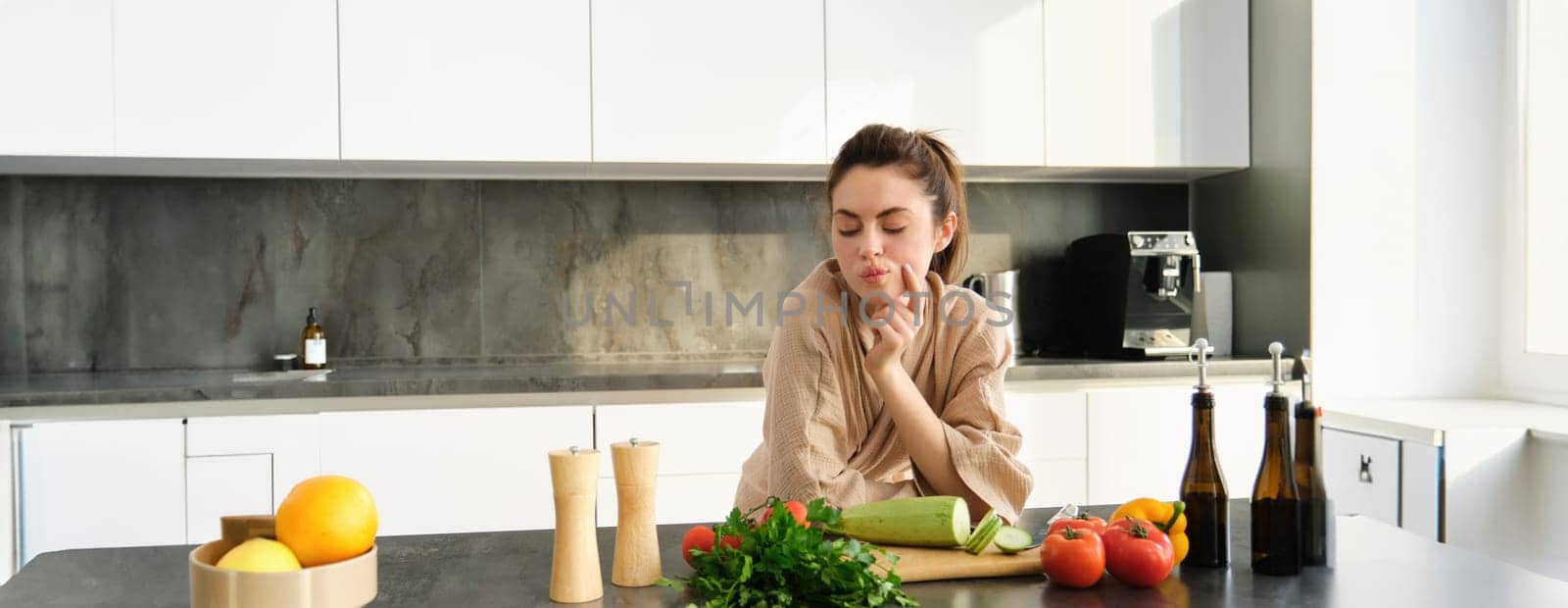 Portrait of woman thinks what to cook with vegetables, looking at chopping board with tomatoes with thoughtful face, standing in kitchen preparing food by Benzoix