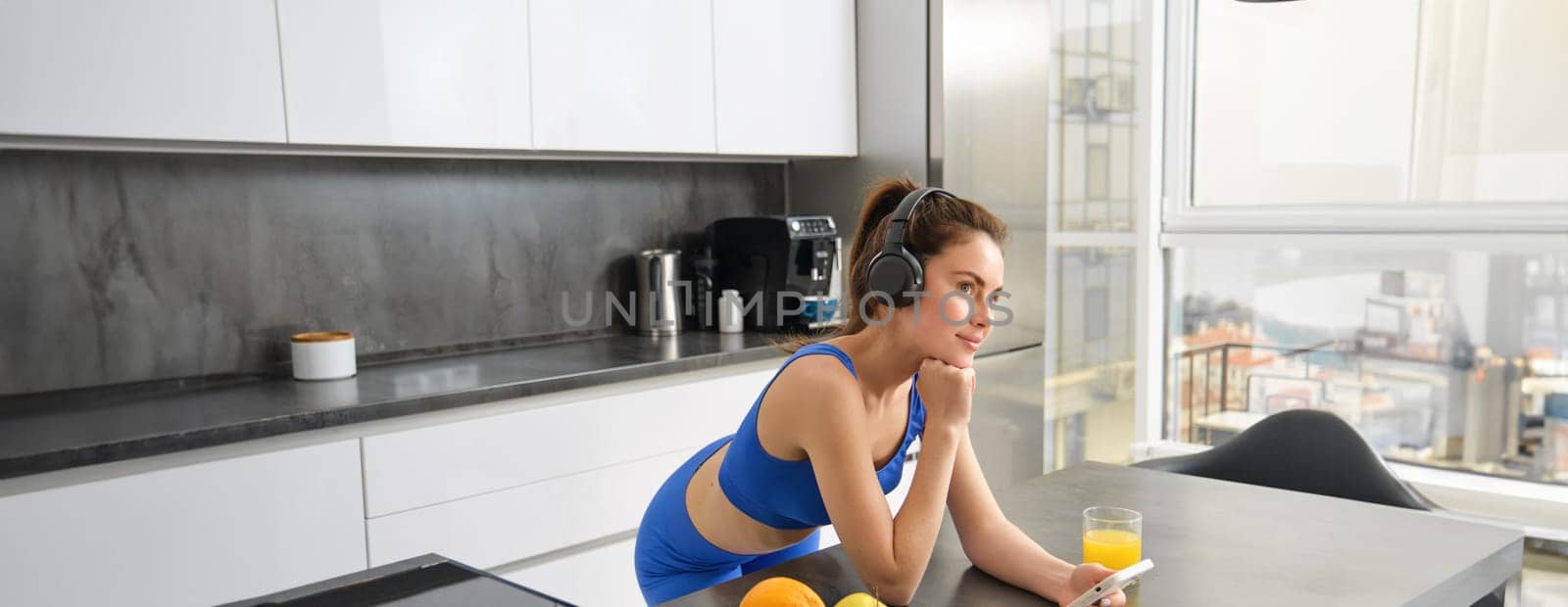 Portrait of stylish sportswoman, listening music in headphones, looking at smartphone, watching video on mobile phone, standing in kitchen, wearing activewear.