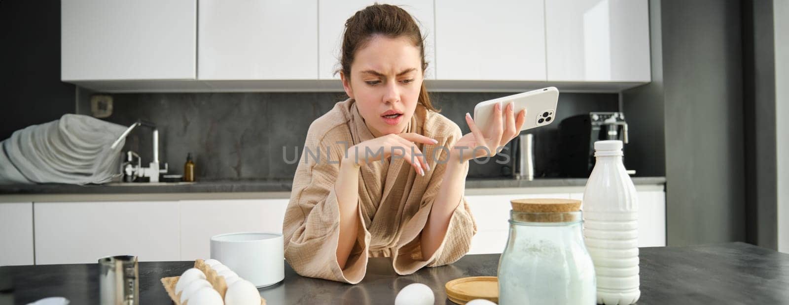 Attractive young cheerful girl baking at the kitchen, making dough, holding recipe book, having ideas.