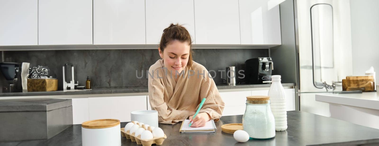 Attractive young cheerful girl baking at the kitchen, making dough, holding recipe book, having ideas.