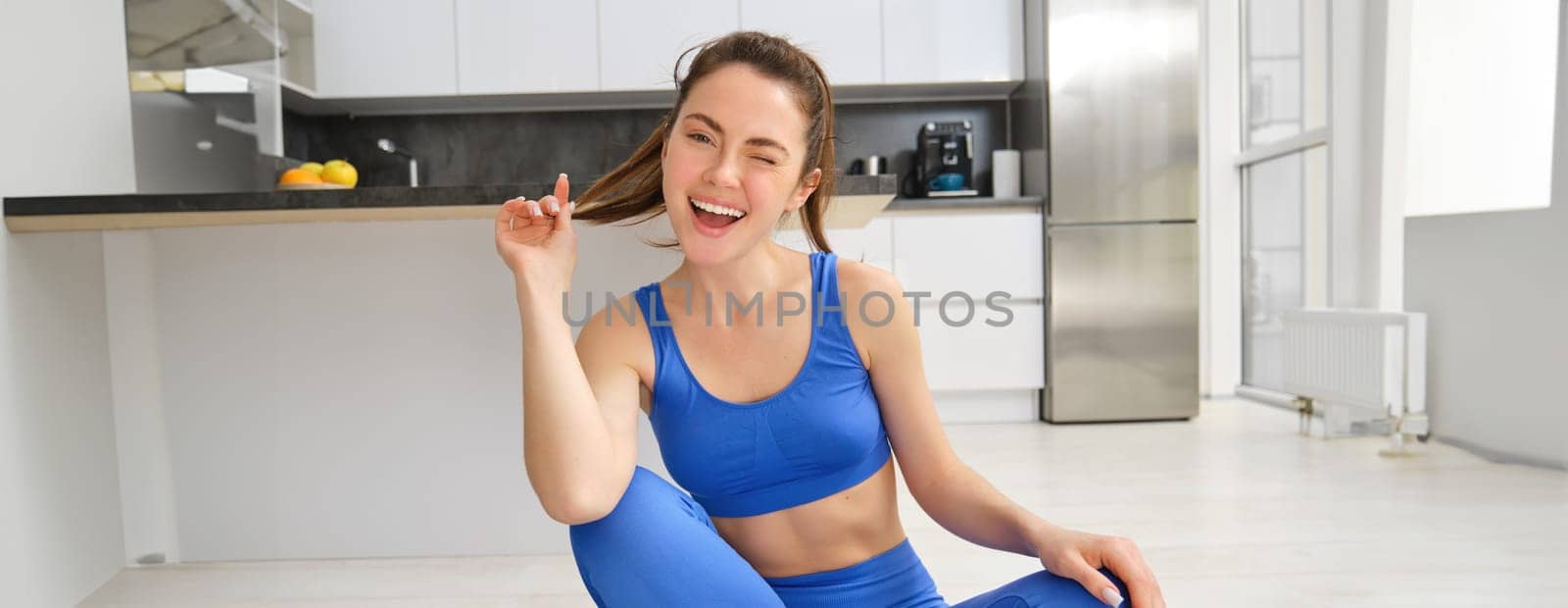 Image of young sport woman sitting at home on yoga mat, doing workout, stretching fitness exercises on floor in living room, smiling and looking happy at camera by Benzoix