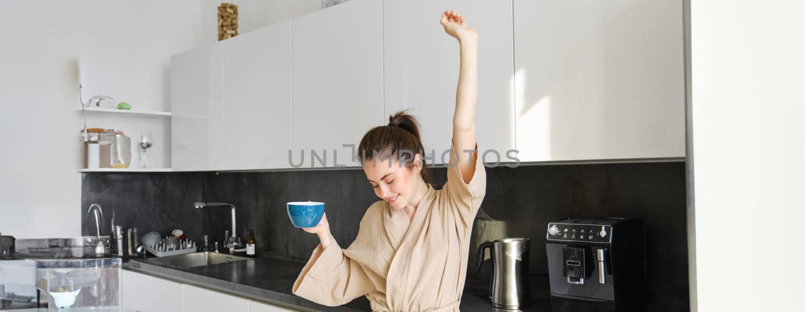 Portrait of happy girl dancing with coffee in the kitchen, wearing bathrobe, enjoying her morning routine.