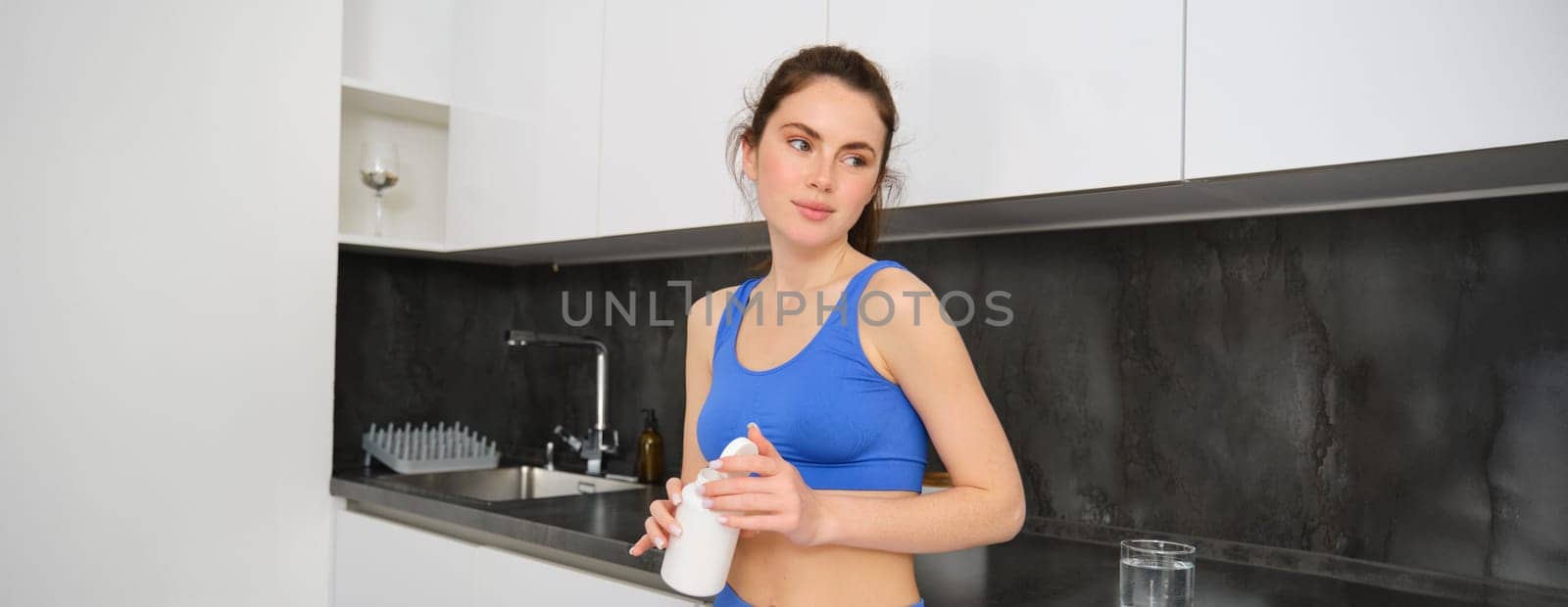 Portrait of healthy, beautiful woman, holding bottle of vitamins, taking dietary supplements, buds for shiny skin and strong muscles, standing in fitness clothing.