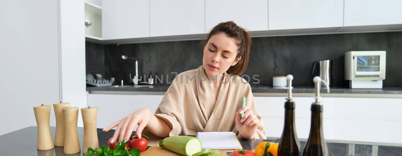 Portrait of woman cooking in the kitchen, sitting in front of vegetables, tomatoes zucchini and parsley, making list of groceries, writing down recipe, wearing bathrobe.