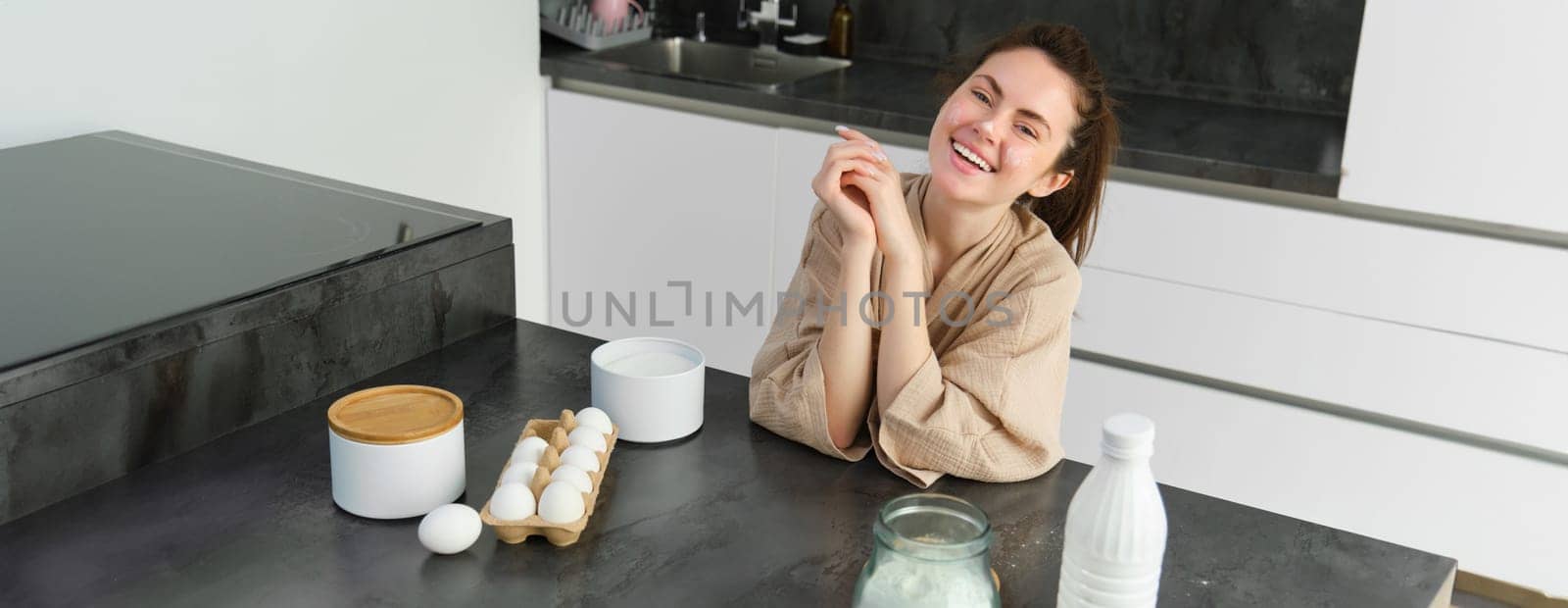 Attractive young cheerful girl baking at the kitchen, making dough, holding recipe book, having ideas.