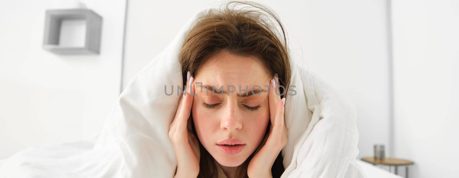 Close up of woman feeling unwell at home, lying in bed under white blankets and frowning, touching head, has headache, migraine, recovering from covid.
