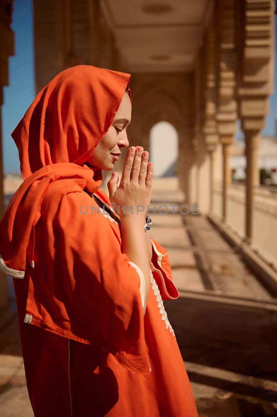 Side portrait of beautiful Middle Eastern Muslim woman in hijab and authentic dress, praying with her palms together while standing between the columns of a beautiful mosque. Photo with perspective