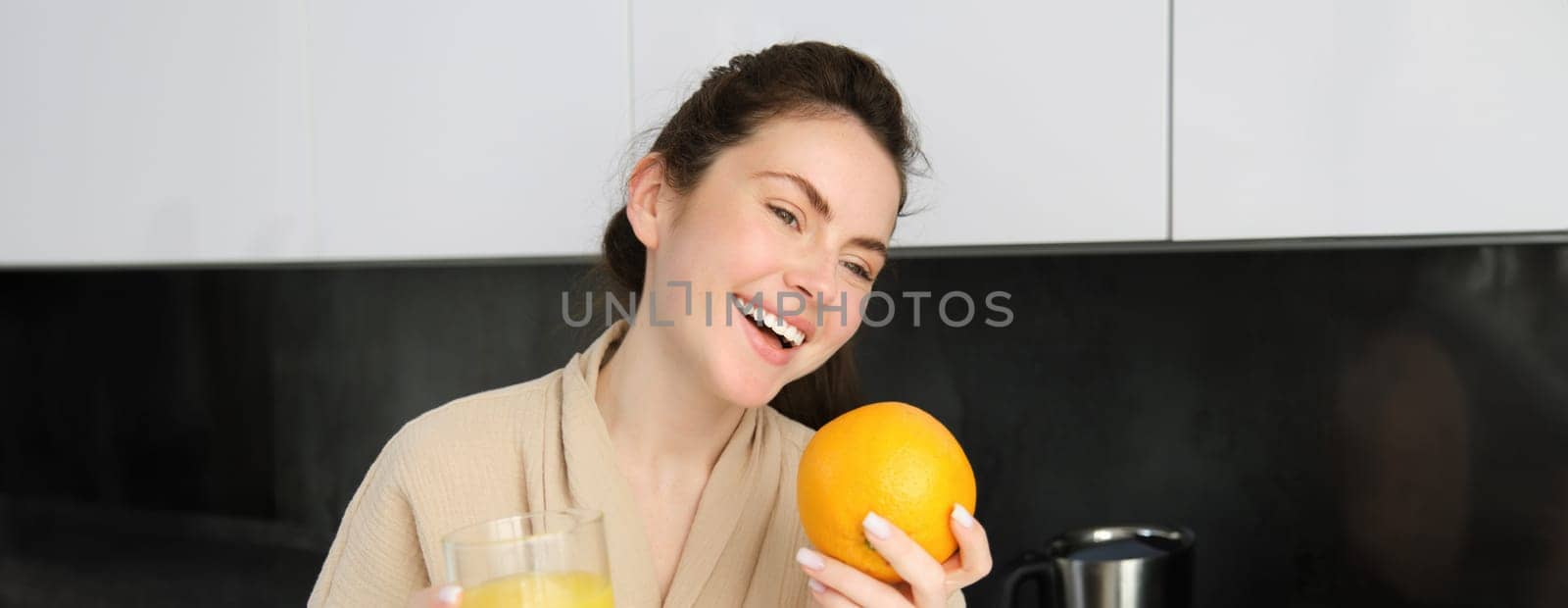 Portrait of attractive young modern woman, posing in kitchen, holding glass of juice and an orange, laughing and talking to someone aside.