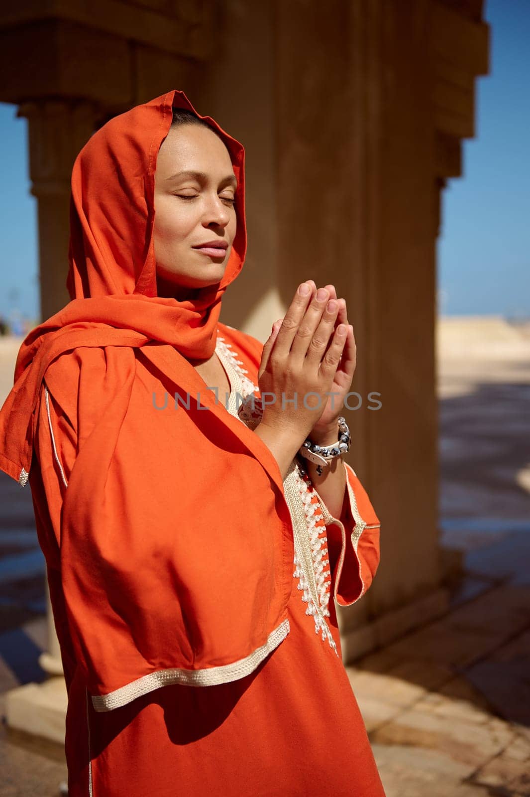 Portrait of beautiful serene Arabian Muslim woman in bright orange authentic dress and her head covered in hijab, praying with her eyes closed, hands up and palms together. People and religion concept
