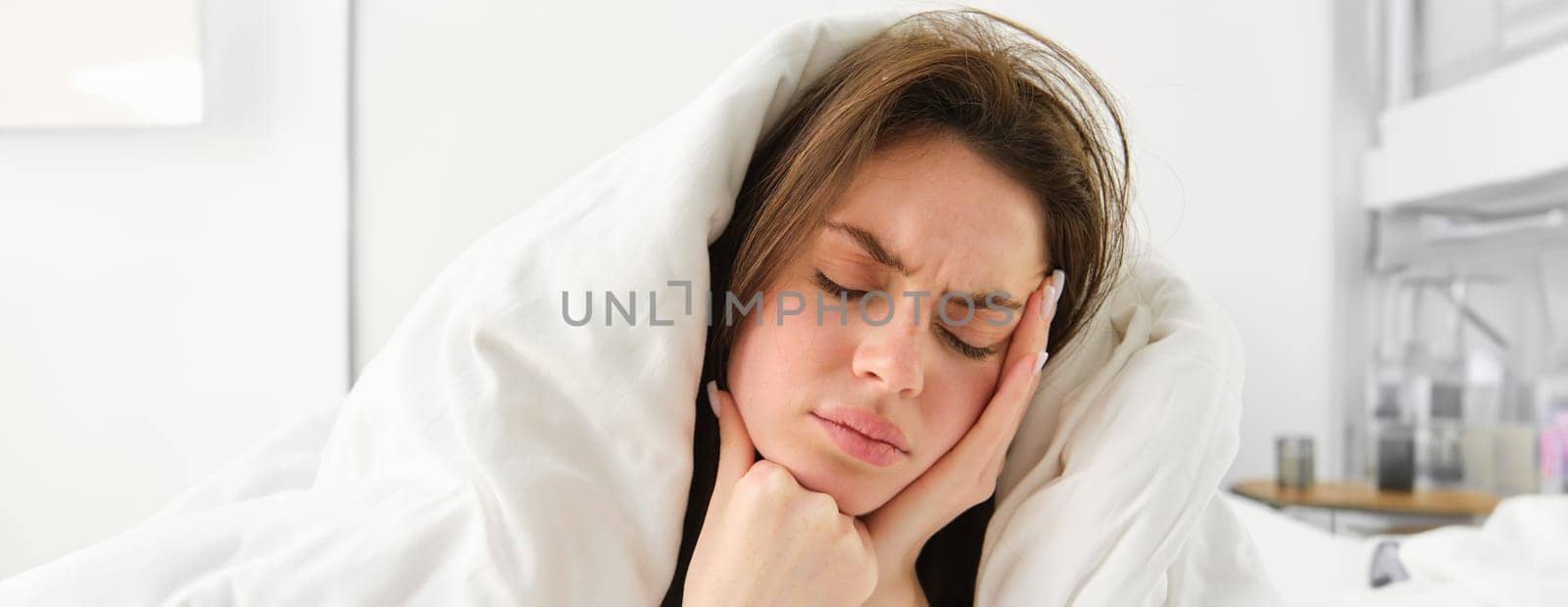 Woman feeling unwell in bed, lying in her bedroom under blanket and white sheets in morning, frowning and touching head, has headache or migraine.