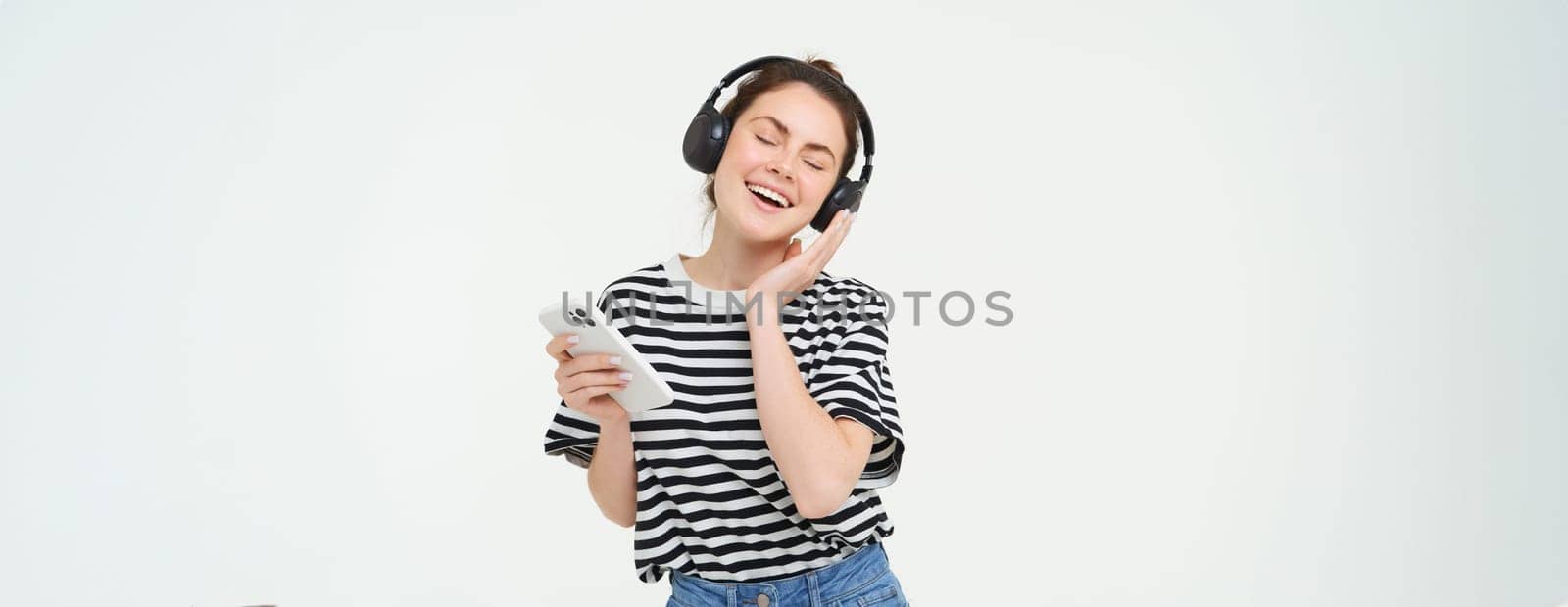 Young woman with smartphone listening to music, dancing to her favourite song in headphones, posing against white background.