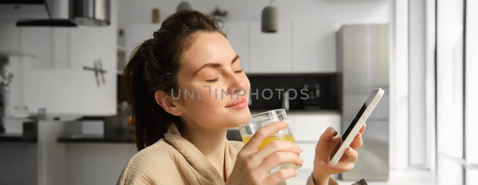 Close up of beautiful smiling woman, drinks fresh orange juice, enjoys her morning, holds mobile phone.