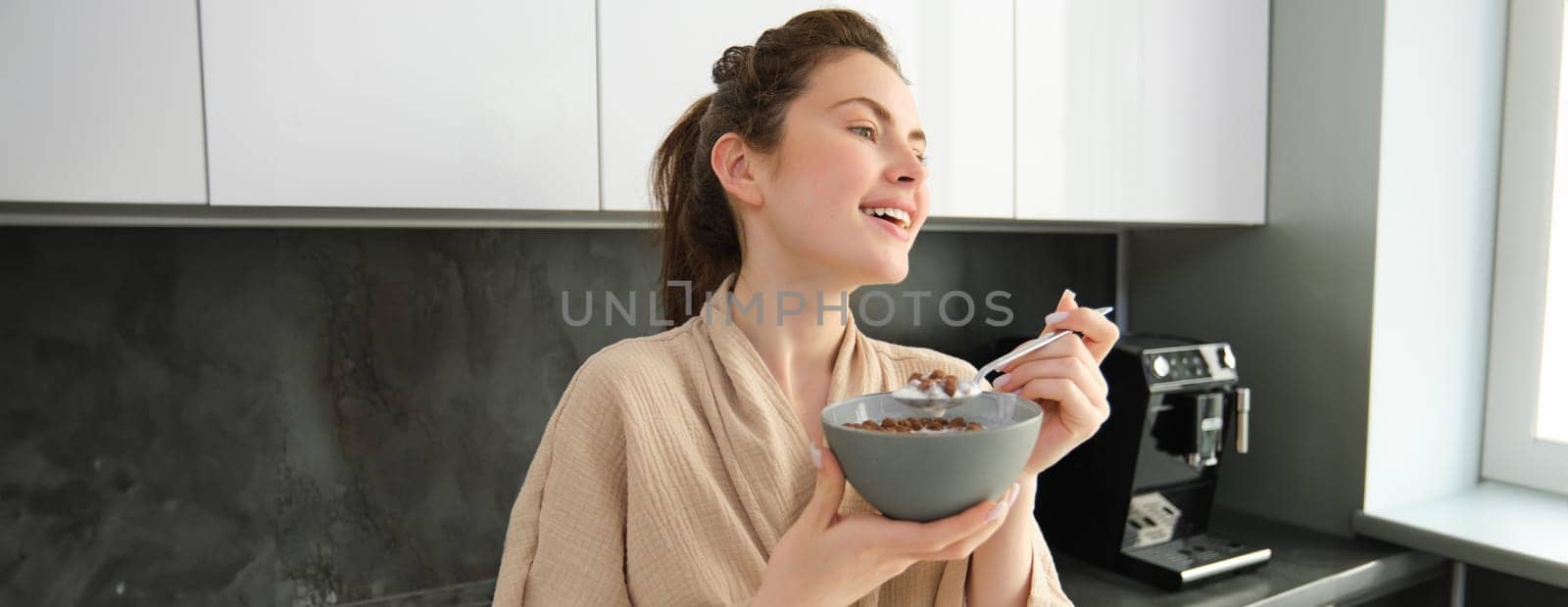 Good-looking brunette woman eating her breakfast, standing in kitchen near worktop and holding bowl of cereals with milk, enjoying her morning, wearing cosy bathrobe.