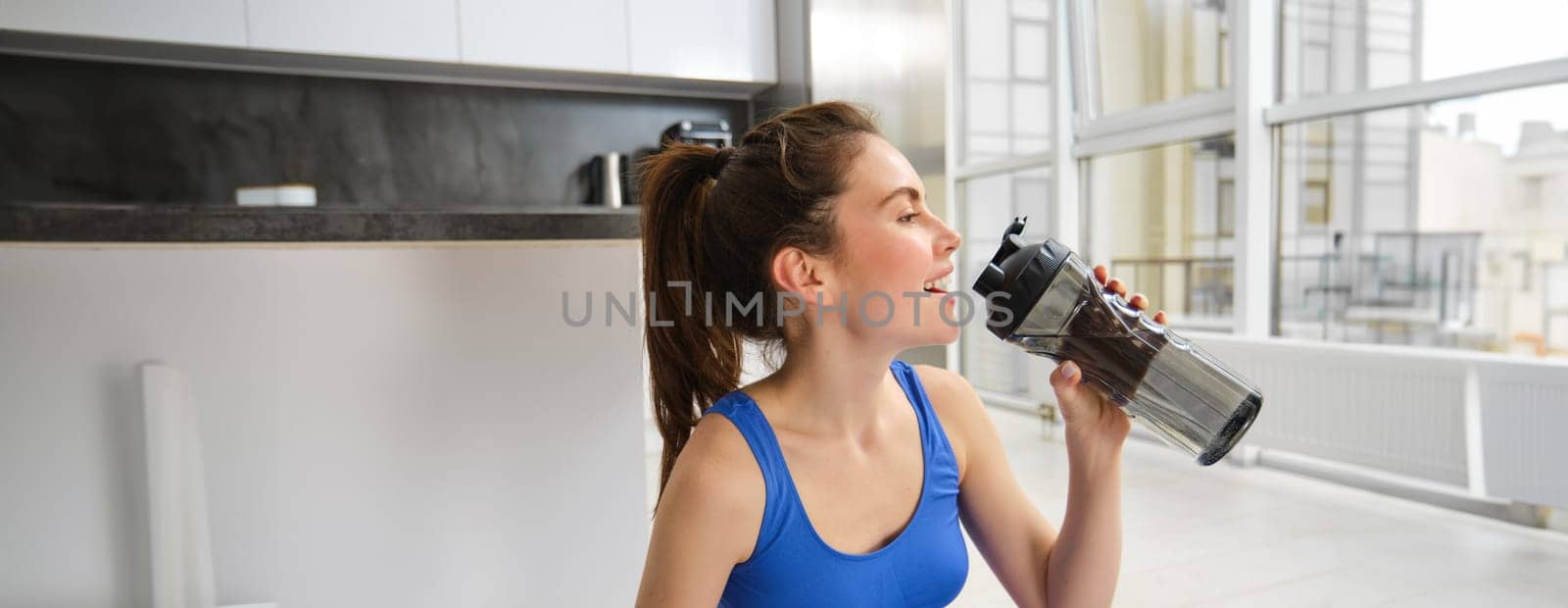 Portrait of sportswoman drinking water during workout session in living room, does her fitness training exercises at home.