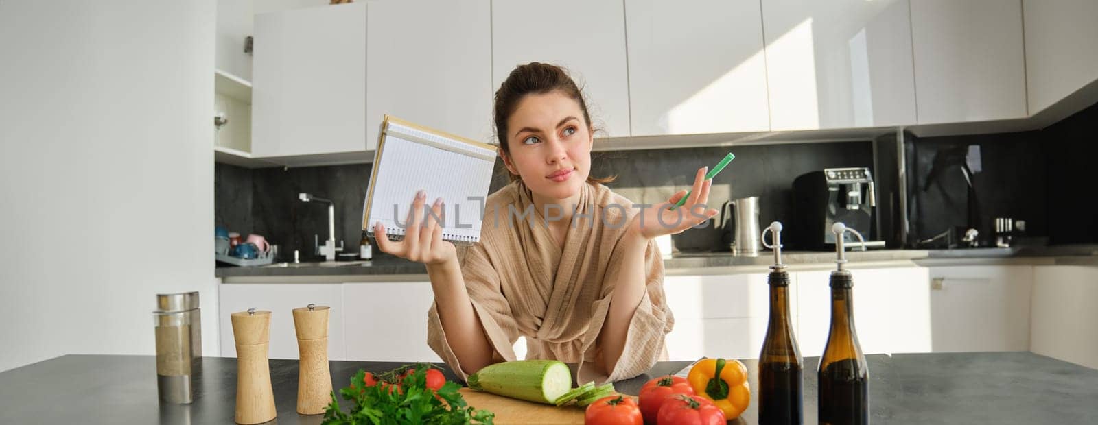 Portrait of woman checking grocery list, looking at vegetables, holding notebook, reading recipe while cooking meal in the kitchen, chopping tomatoes and zucchini.