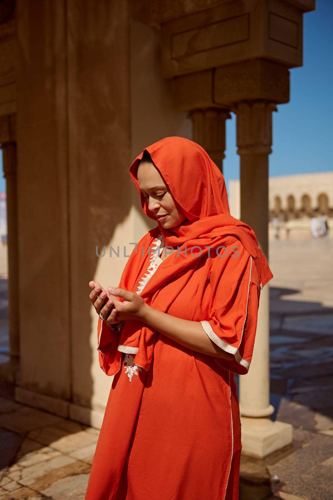 Portrait of young Arabic Muslim woman praying standing near marble columns at mosque. Religious lady with her eyes closed, in bright orange head scarf making Islamic pray Dua gesture with cupped hands