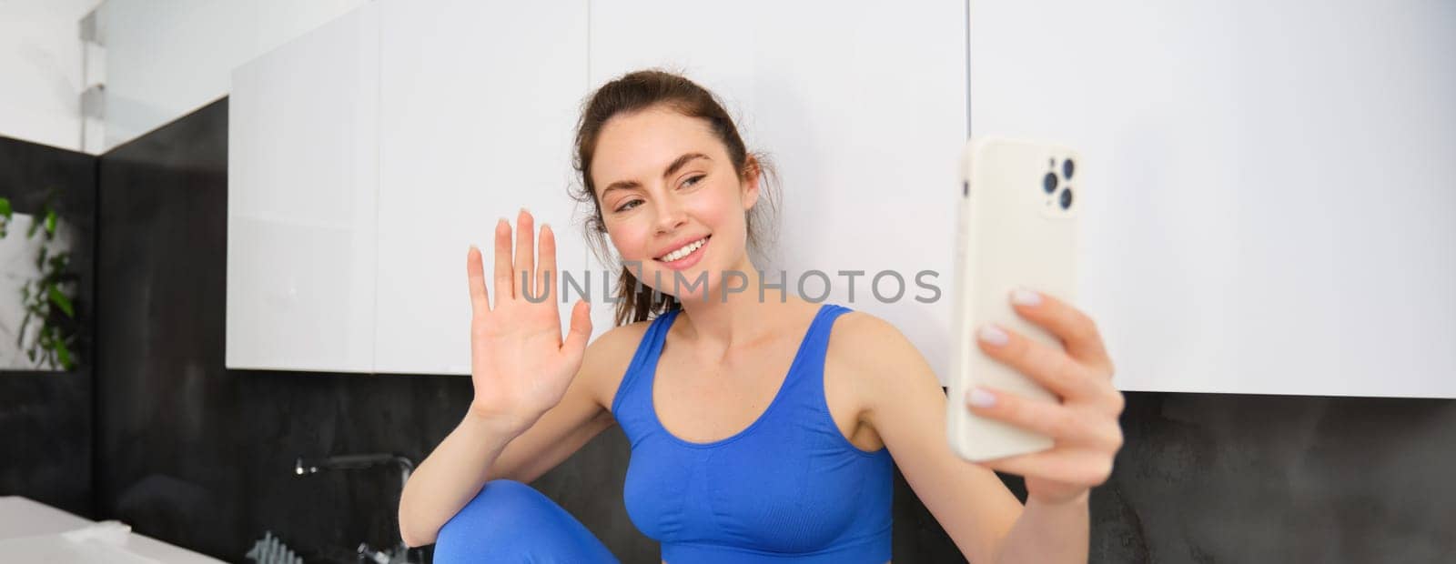 Portrait of young fitness girl, social media blogger, live streaming, saying hello at smartphone, waving hand and smiling, wearing activewear by Benzoix