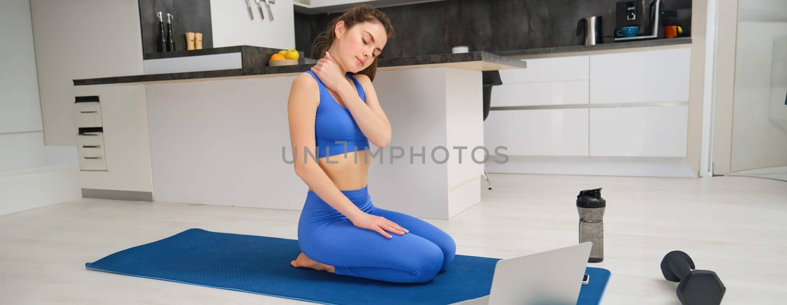 Image of calm and relaxed woman, sitting on yoga mat with laptop, massaging her neck, warm-up body before workout training, home fitness session by Benzoix