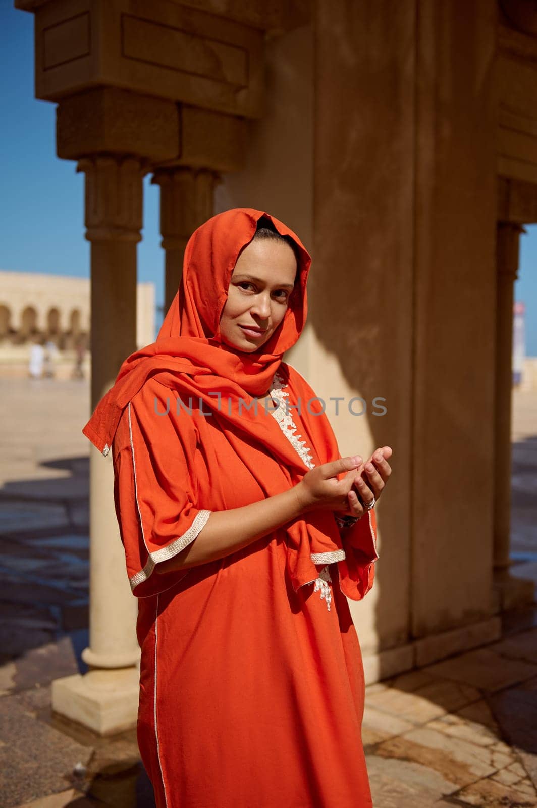 Beautiful Middle Eastern Muslim woman with head covered in hijab and bright orange authentic traditional wear, stands with her hands palms together, looks at camera while walking at Hassan II mosque