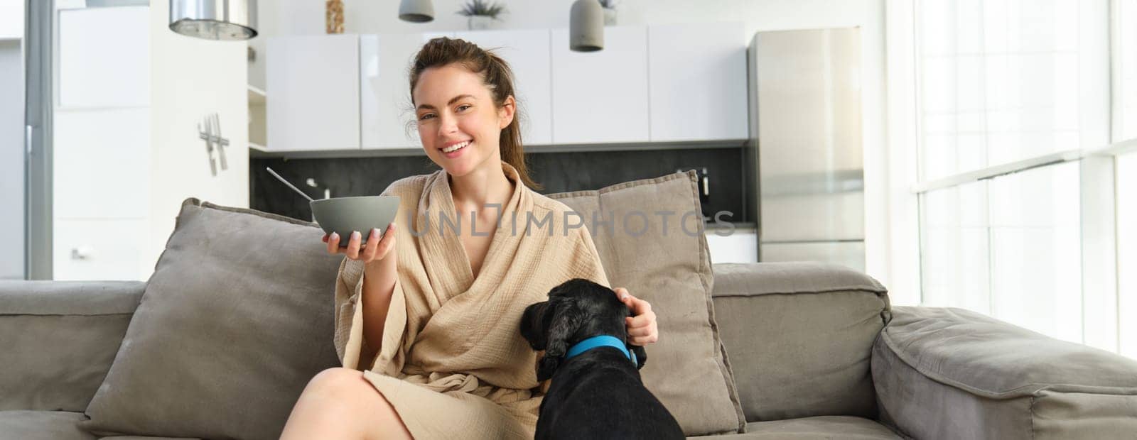 Happy mornings. Beautiful woman enjoying her morning, sitting on sofa, eating breakfast and playing, cuddling with dog, puppy asks for small bite of cereals.