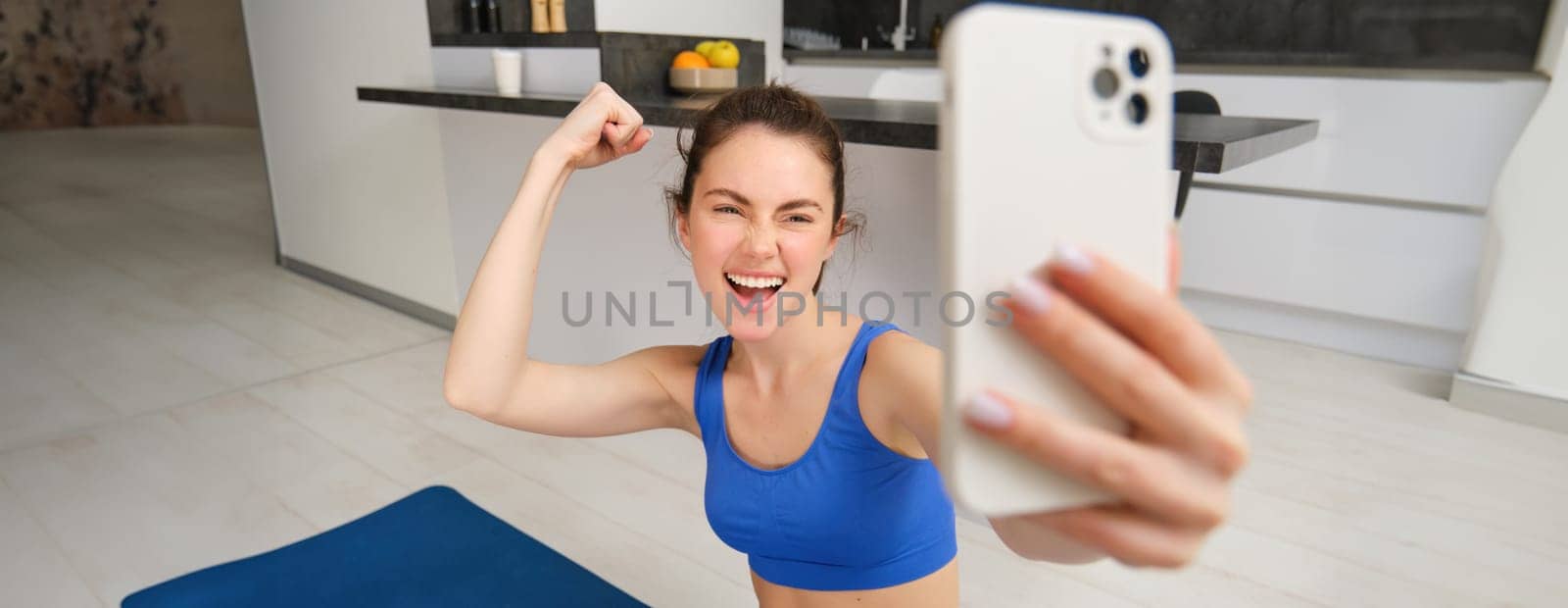 Young energetic girl does sports at home, shows her muscles on arm and takes selfie on smartphone, posing on fitness rubber mat in living room by Benzoix