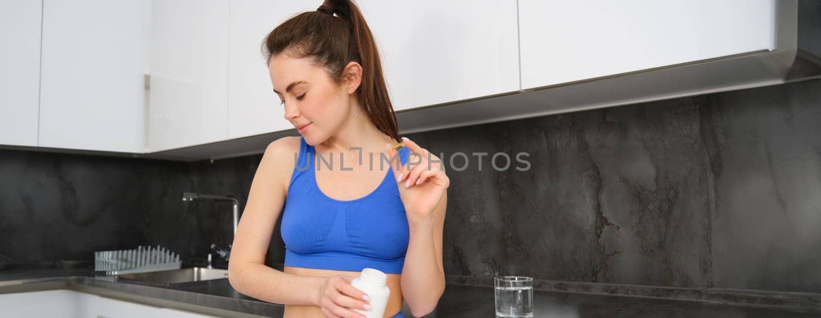 Image of young brunette woman with slim, fit body, holding dietary supplements, vitamin B, fish oil, standing in kitchen by Benzoix