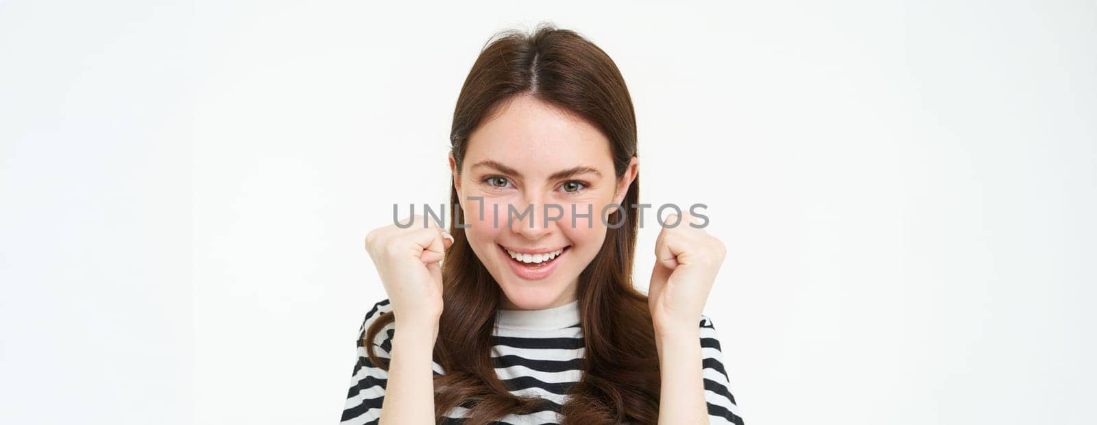 Image of young woman looking with pleased, satisfied face at camera, winning, shaking hands in triumph, celebrating victory, achievement, standing over white background.