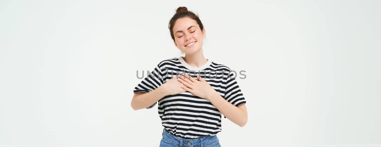Portrait of happy, beautiful brunette woman, holds hands on heart and smiles, stands in casual clothes over white background. Copy space