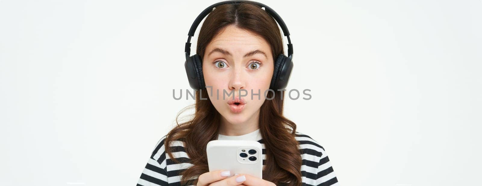 Portrait of happy young modern woman, female model in wireless headphones, smiling and looking at camera, holding smartphone, using mobile phone app, white background by Benzoix