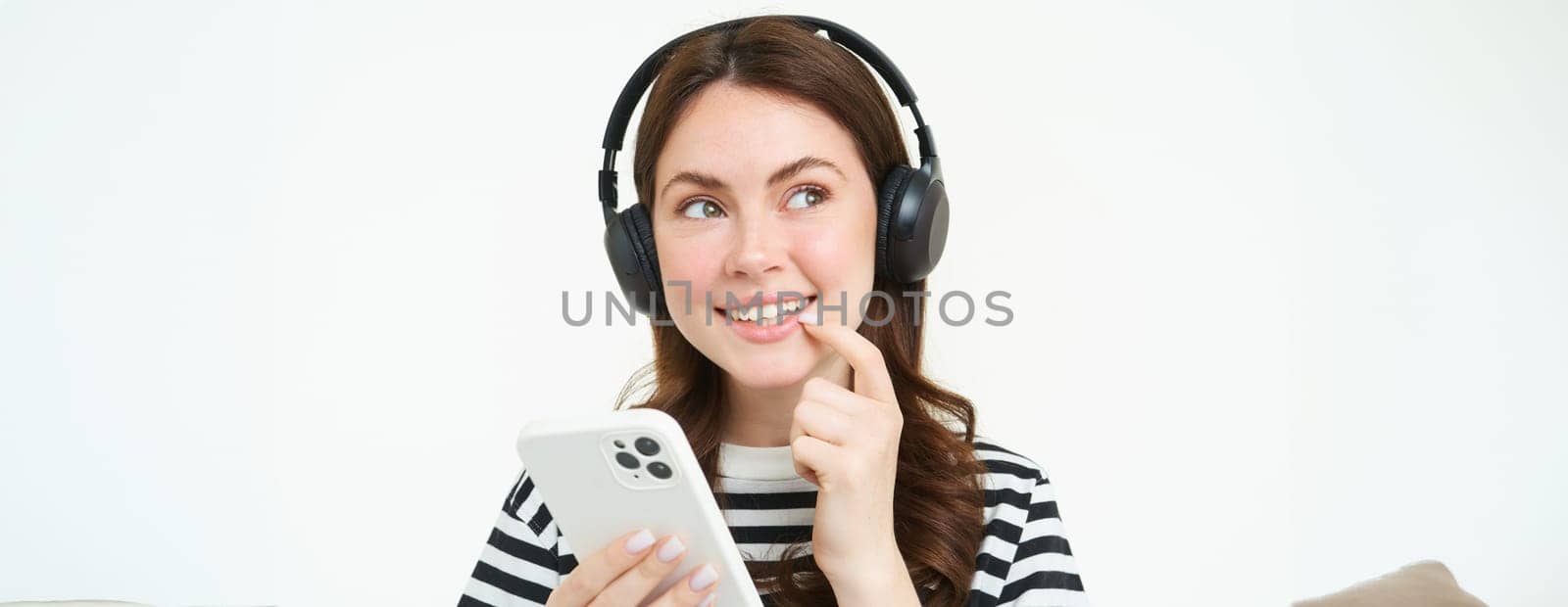 Image of girl in headphones, smiling while thinking, making decision, holding smartphone, making choice, online shopping, white background.