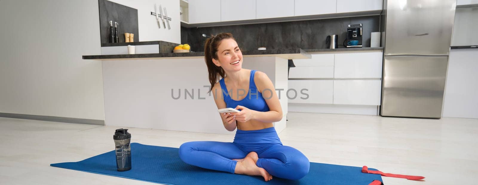 Portrait of woman using fitness app, doing workout from home, sits on yoga mat with smartphone in living room.