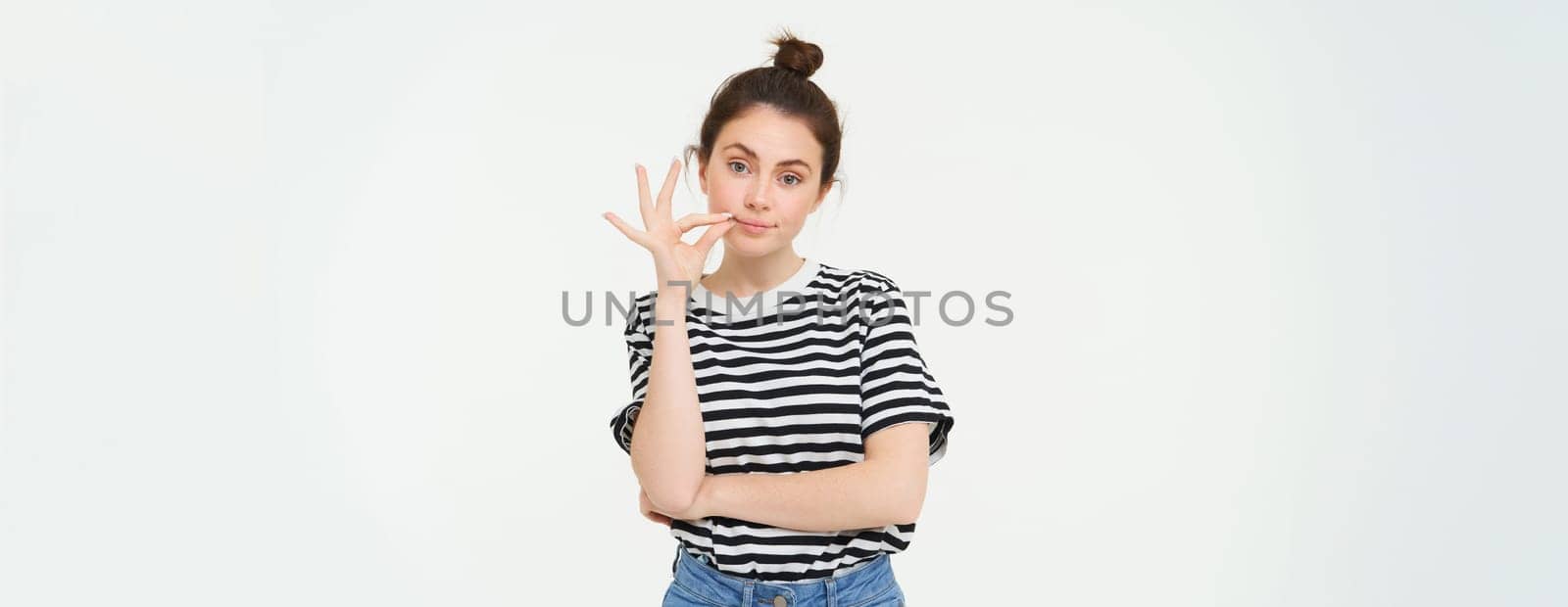 Portrait of brunette woman seals her mouth with promise not to tell anyone, zipping gesture, standing over white background.