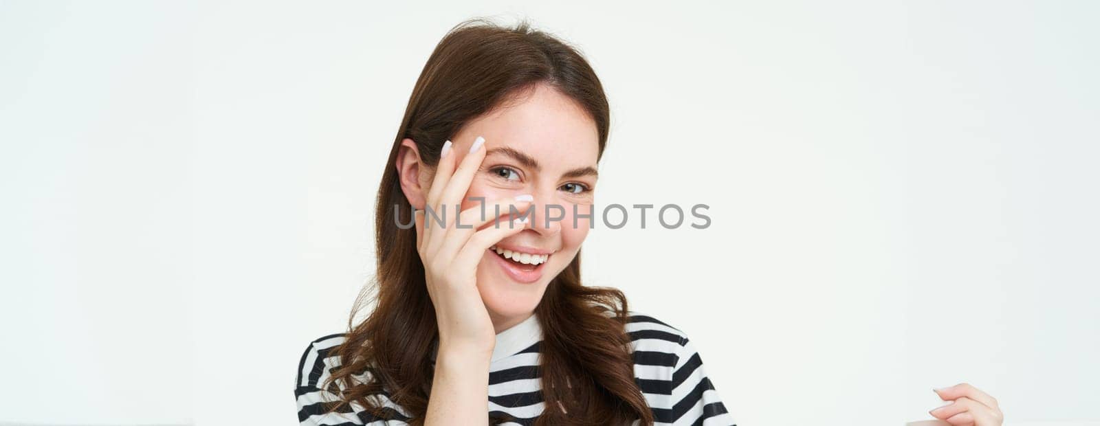 Portrait of attractive, smiling young woman, laughing and smiling, touches her face, standing over white background.